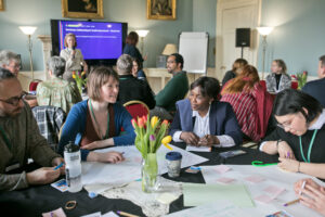 Groups of adults sit around circular tables and engage in discussion. The tables are covered with large sheets of paper, post-it notes, and pens.