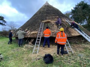 A group of people wearing hard hats and hi vis jackets in front of and on top of a wooden roundhouse with a large thatched roof.