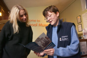 A museum staff member with light skin tone, short brown hair, and glasses is standing on the right/ On the left is an adult with light skin tone and long blonde hair. They are both looking at a booklet that says 'Ullapool Museum' across the front.