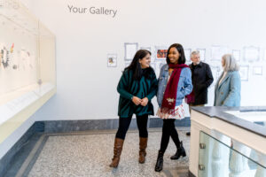 Four adults walking through a bright gallery space with white walls and marble floors. The person at the front on the left has long dark hair and medium skin tone, the person at the front on the right has short dark hair and medium to dark skin tone. The people walking behind have short grey hair and light skin tone.