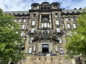Front-facing view of Glasgow Royal Infirmary Museum which is a large stone building built in the early 1900s. The building features tall columns, decorative carvings, and many rows of windows. A bronze statue of Queen Victoria sits above the entrance.