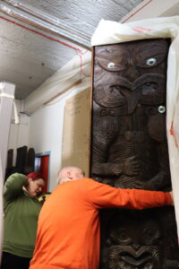 A person with pink hair wearing a green jumper and a person with a close shaven head wearing an orange examine an ornately carved dark wooden Māori poupou panel with iridescent shells attached to the wood.