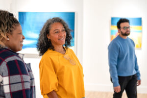 An adult with medium-dark skin and a brown wavy ponytail, and adult with medium-tone skin and curly grey-black hair, and an adult with medium-tone skin, short black hair and a beard stand in a gallery with white walls and brightly-coloured abstract paintings.
