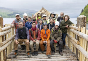 A group of adults in hard hats and hi-vis vests posing for a photo on a wooden walkway that leads to a wood roundhouse.