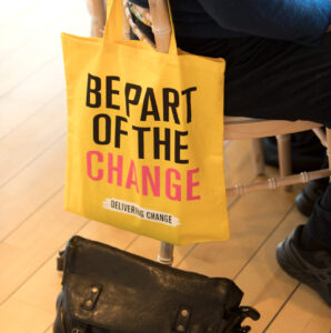 A bright yellow canvas tote bag hangs from the back of a wooden chair. The bag has the words "BE PART OF THE CHANGE Delivering Change" printed on the front.