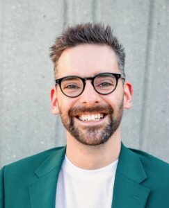 An adult with light skin, short grey-brown hair, a short beard, and glasses.