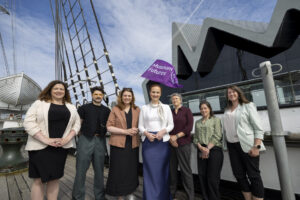A group of seven adults are standing aboard a ship. A large mast is standing tall behind them with a purple flag attached that reads 'Museum Futures'. A large grey building with an angular roof is also in the background.