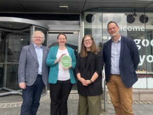 Four adults stand together and smile in front of a building with glass walls and a revolving door. One member of the group - and adult with light skin, tied-back brown hair, and a green jacket - holds a green plaque which features the text "Green Tourism".