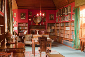 A grand red room with bookcases lining the walls and art hanging above them. A light fixture hangs above an arm chair in the centre of the room behind a desk. A fire place sits on the back wall with a round mirror above it.