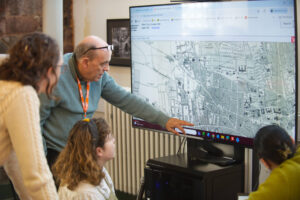 An adult with light skin tone and short grey hair. They are wearing a green knitted jumper and orange lanyard. They are pointing at a large screen with a black and white planning map of Dundee displayed on it.