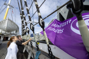 Two adults hoist a large purple flag bearing the text "Museum Futures" up from the rigging of a ship.
