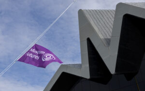 A purple flag with white writing that reads 'Museum Futures'. A grey building with an angular roof is in the background.