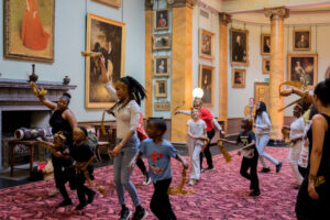 Children and adults with light brown and dark brown skin dancing and running through a room with high ceilings, marble columns and framed portrait paintings of people with pale skin.