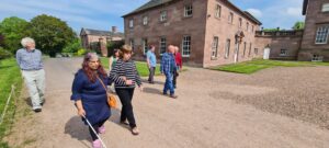 A woman with brown, long hair is using a white cane and linking arms with another woman in the foreground. several other people are walking nearby in front of a large manor house.