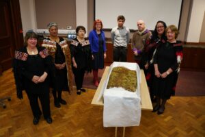A group of people, some with darker skin tone, others with lighter skin tone, stand around a Kahu Kākāpō feathered cloak lying on a white table cloth. Many of the people in group are wearing ceremonial cloaks.