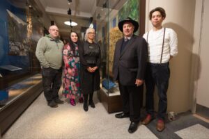 A group of people pose in front of a museum glass case containing a feathered Kahu Kākāpō cloak.