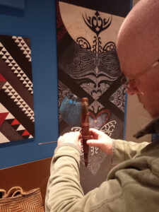 Looking over their shoulder, a person with light skin, glasses and a bald head attaches a wooden musical instrument to a fixture in a glass case.