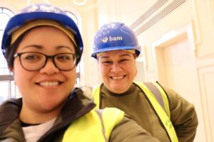 Two people with olive skin tone, wearing blue hard hats and high vis vests, take a selfie together, smiling towards the camera.