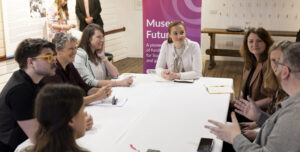 Eight adults are sitting at a large white table in discussion. The person at the head of the table is Culture Secretary, Màiri McAllan who has light skin tone and dark red hair tied up.