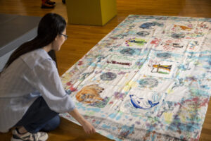 A person with light skin tone and long dark hair is crouching over a large white cloth covered in colourful paintings.
