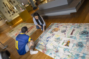 Two adults are crouched over a large cloth which is laid out on a hard wood floor. The cloth has a white background with many colourful images painted across it.