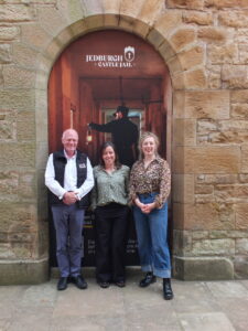 Three adults standing in front of an arch which features a poster promoting Jedburgh Castle Jail.