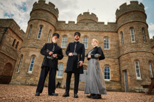Three adults are standing together dressed in prison guard outfits from the Victorian period. The two on the left are wearing black coats and trousers with flat topped black hats. The person on the right is wearing a grey dress and black coat. Behind them is a two storey, 19th century building with large arched windows.