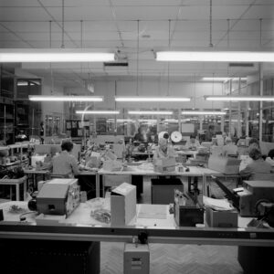 A black and white image of a watch factory room filled with people sitting at desks. The people are assembling watches by hand.