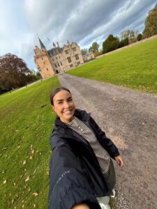 A young woman with brown hair tied back takes her own photo at the end of a long driveway with a castle in the background.