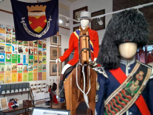 A gallery space containing a mannequin in a red jacket and grey top hat, sitting on a wooden horse and holding a blue flag which features a coat of arms. On the wall behind the mannequin is a display of colourful brochure covers.