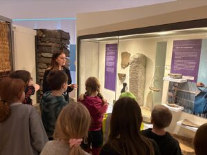 A woman with brown hair stands in front of a museum display case, speaking to a group of children gathered around her.