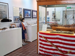 A gallery space with white walls and a red tiled floor. Display cases containing naval artefacts line the walls. In the middle of the room is a large wooden display case containing a model sail ship. On the side of the case is a red and white striped flag which features an image of a snake.
