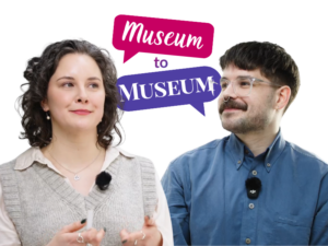 An adult with light skin and wavy grey-black shoulder-length hair engages in conversation with an adult with light skin, short brown hair, a moustache, and glasses. Behind them is a graphic of two speech bubbles containing the text: "Museum to Museum".