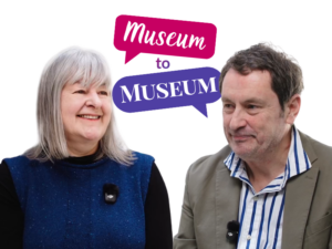 An adult with light skin and straight grey shoulder-length hair engages in conversation with an adult with light skin, show brown hair, and a grey stubble. Behind them is a graphic of two speech bubbles containing the text: "Museum to Museum".
