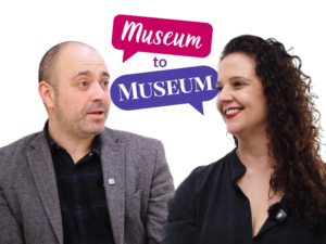 An adult with medium-light skin, short black hair, and a stubble engages in conversation with an adult with medium-light skin and long curly dark brown hair. Behind them is a graphic of two speech bubbles containing the text: "Museum to Museum".