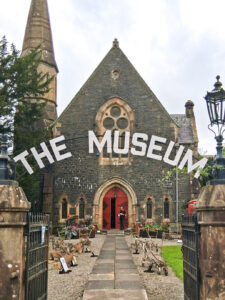 The front of a church building with grey stone walls, gothic windows, and a large red door. In the foreground is a gateway with the words "The Museum" in large white text over the arch.