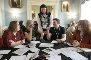 A man with long hair stands at the centre of a round table, with four people seated two on each side, everyone smiling.