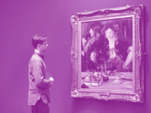 An adult with light skin and short brown hair observes an oil painting of an adult with light skin and black hair seated at a table.