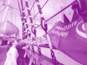 Two adults hoist a large purple flag bearing the text "Museum Futures" up from the rigging of a ship.